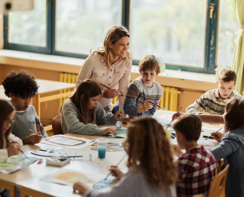 Eine Gruppe von Schüler:innen sitzt um einen großen Tisch und malt. Eine Lehrerin steht mit Pinsel in der Hand und lächelt einer Schülerin zu.