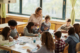 Eine Gruppe von Schüler:innen sitzt um einen großen Tisch und malt. Eine Lehrerin steht mit Pinsel in der Hand und lächelt einer Schülerin zu.