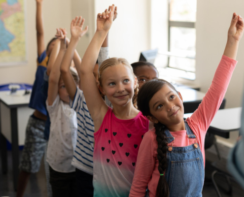 Volksschulkinder stehen in einem Klassenzimmer in einer Reihe hintereinander und heben die Hände in die Höhe