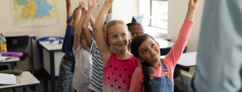 Volksschulkinder stehen in einem Klassenzimmer in einer Reihe hintereinander und heben die Hände in die Höhe