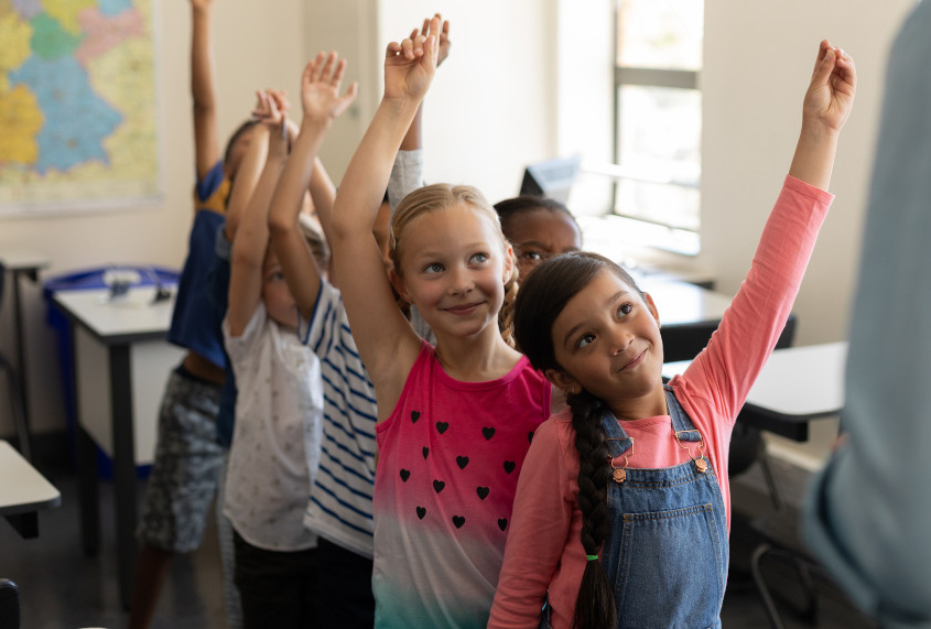 Volksschulkinder stehen in einem Klassenzimmer in einer Reihe hintereinander und heben die Hände in die Höhe